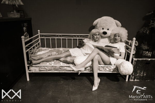 Twee dragqueens en een grote knuffelbeer liggen samen op bed en lezen uit een kinderboek