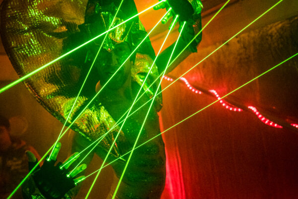 Een foto van een danser met felgroene laserstralen vanuit diens vingers en met rode achtergrondbelichting.